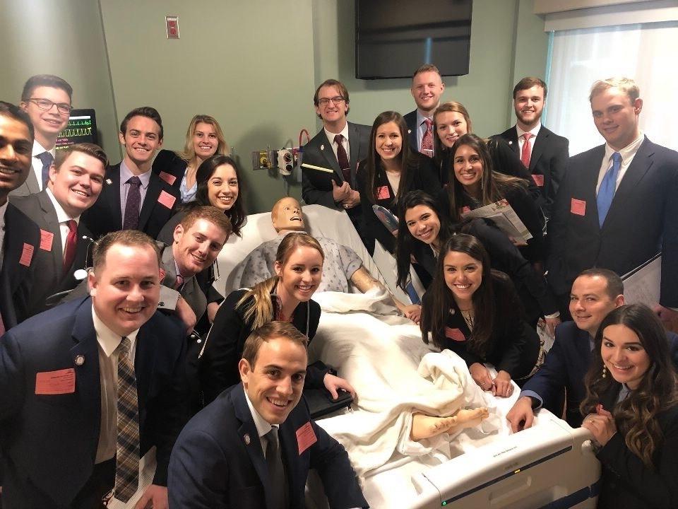 Students pose with mannequin of a patient on a gurney.