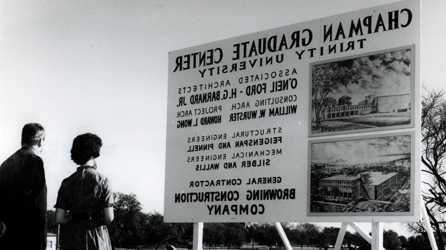 A man and woman looking at a wooden sign promoting the construction of Chapman Graduate Center at Trinity University.